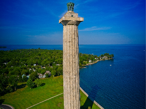 Tall stone monument by the lake with trees and houses nearby.